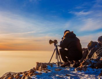 Professional photographer takes photos with camera on tripod on rocky peak at sunset. Dark tone.